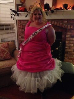 A woman wearing a pink sash and tiara in front of a fireplace on Halloween.