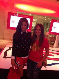 Two women attending a Kansas City Chiefs style party, standing next to each other on a red carpet.