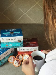 A woman is holding a cup of coffee next to a box of Cameron's k-cups.