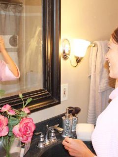 A woman using Olay Facial Cleanser in a pink robe smiling in front of a mirror.