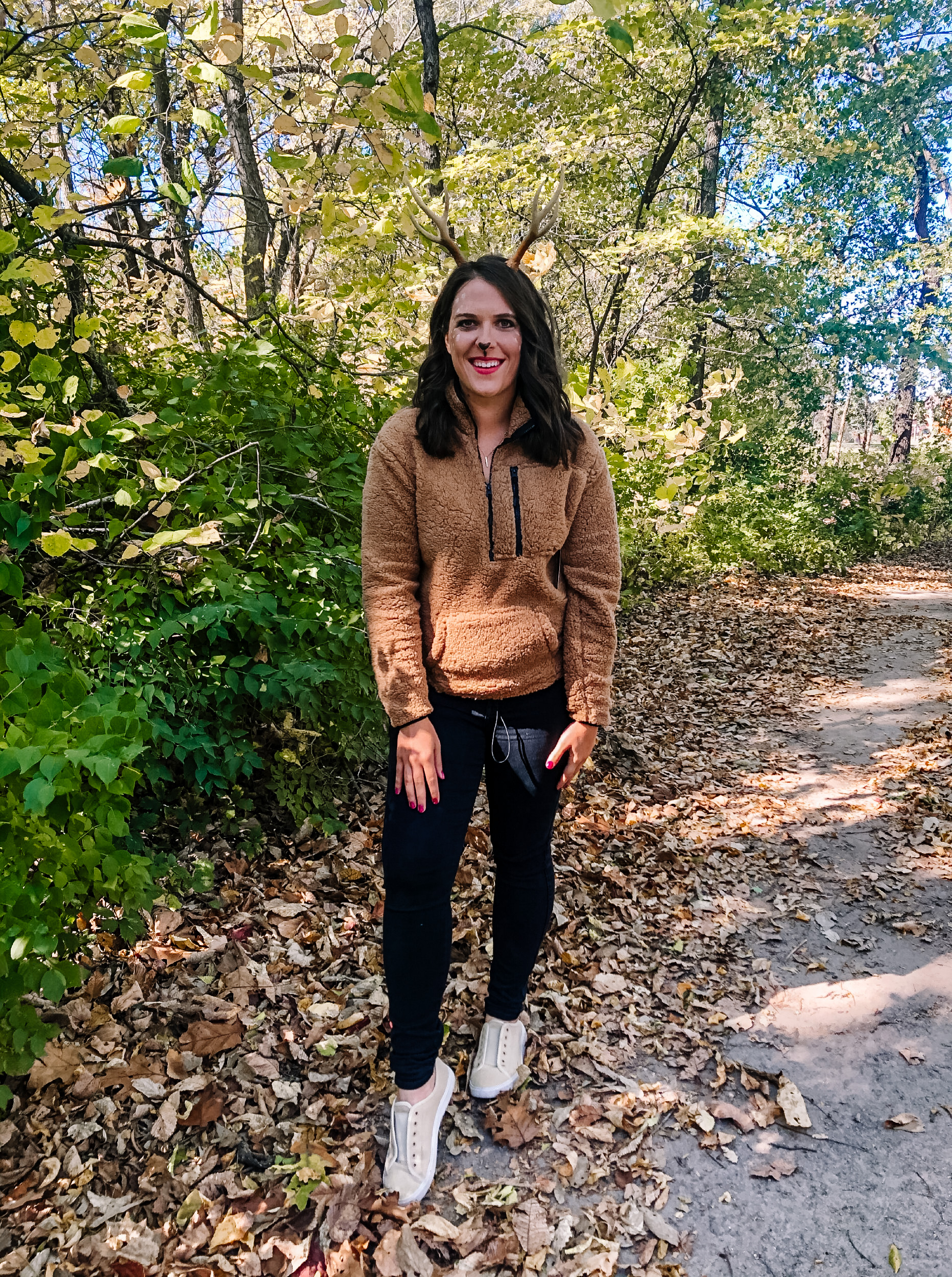A woman wearing a brown sweater and black leggings in the woods, showcasing easy deer makeup for Halloween.