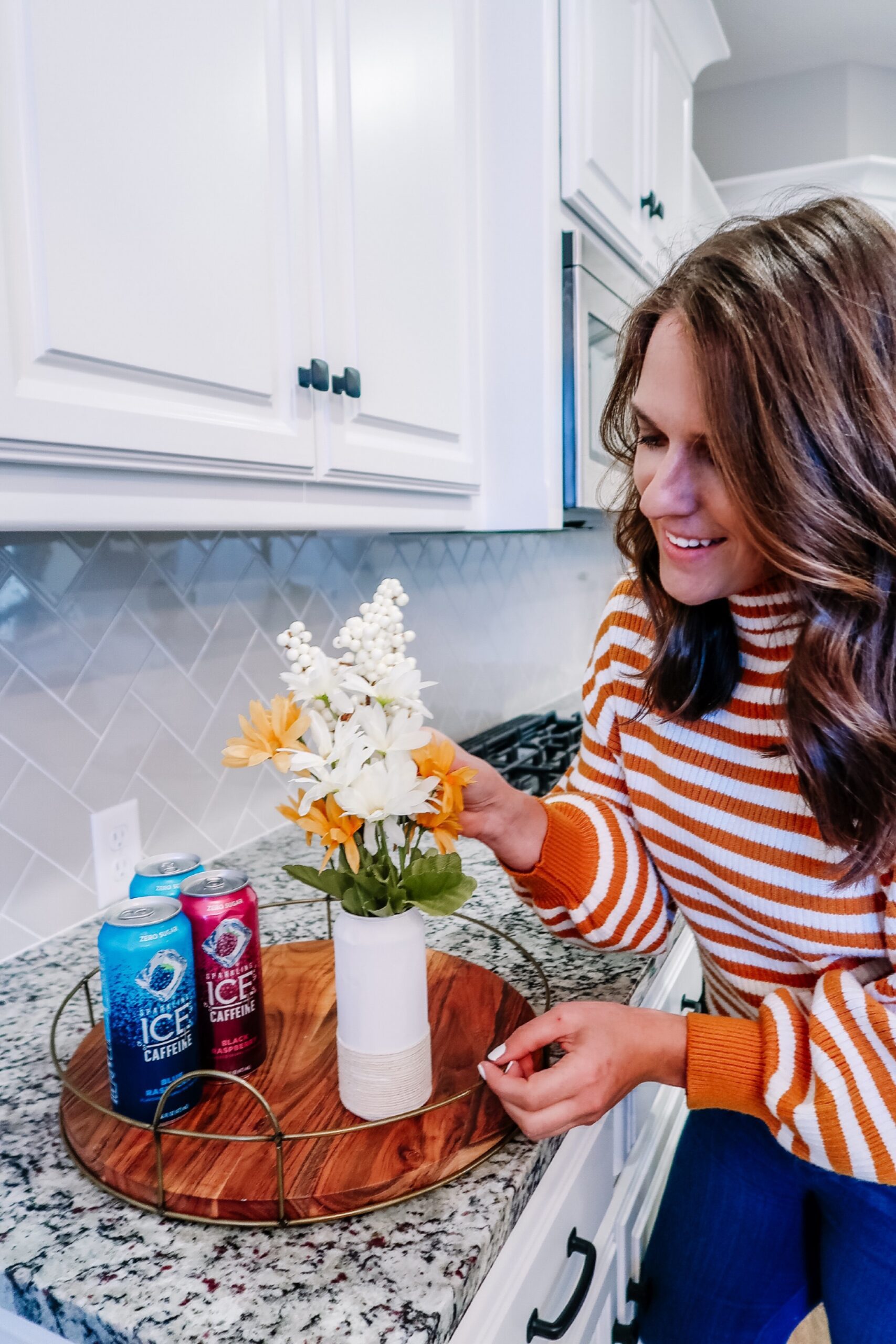 A woman is creating a DIY holiday centerpiece by arranging flowers in a vase in the kitchen.