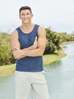 A man, possibly Becca's potential bachelor, in a blue tank top and shorts standing next to a river.