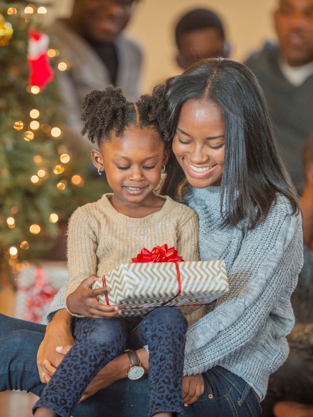 a girl holding a christmas present is sitting with her mom in front of the christmas tree