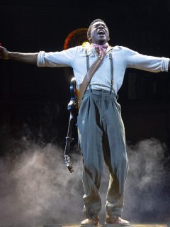 A man on stage with his arms outstretched in the Hadestown musical.