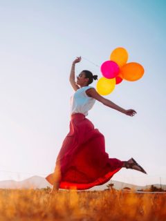 A woman is running with colorful balloons in a vibrant field, embracing the beauty of manifestation.