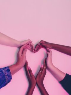 A group of hands making a heart shape on a pink background, representing the manifestation of love and affirmations.
