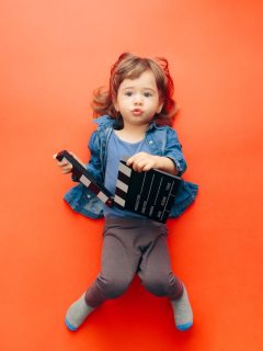 A little girl holding a movie clapper on a red background, aspiring to become a Disney Channel actress.