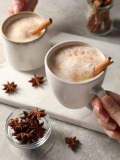 Two hands holding a cup of coffee with cinnamon and star anise.
