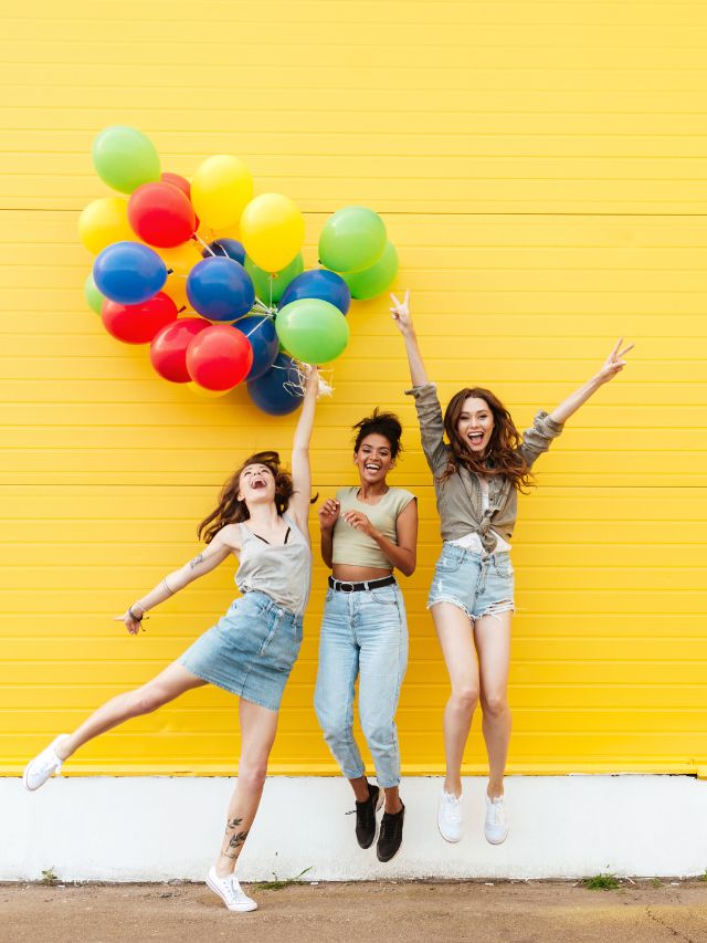 Three young women jumping with colorful balloons against a yellow wall.