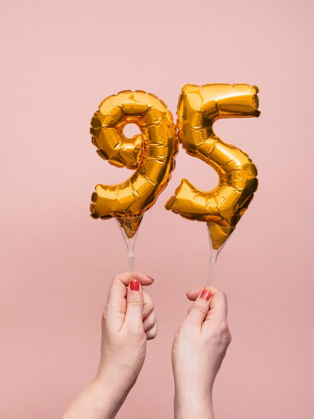Two hands holding gold foil balloons with the number 95 on them.