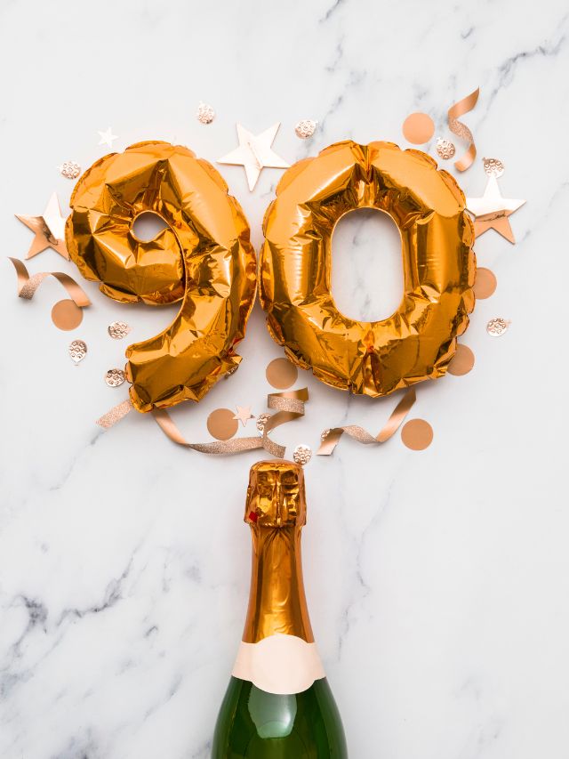 A bottle of champagne and gold foil balloons on a marble table, symbolizing the angel number 90.