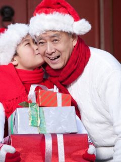 A man in santa hat kisses his daughter while holding Christmas presents.