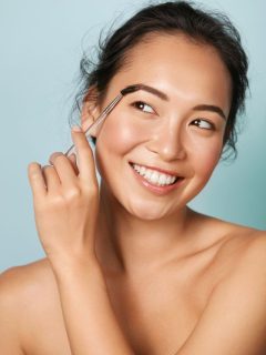 Asian woman applying makeup with a brush on blue background.