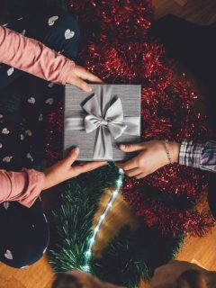 Two people are holding a gift box in front of a Christmas tree, showcasing the best gender-neutral Christmas gifts.