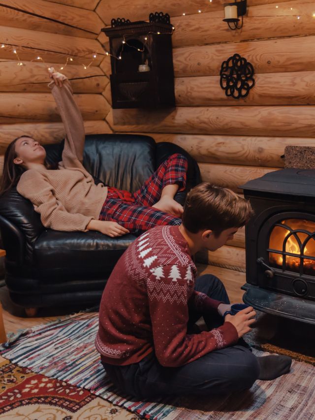Two people sitting in front of a wood burning stove in a log cabin.