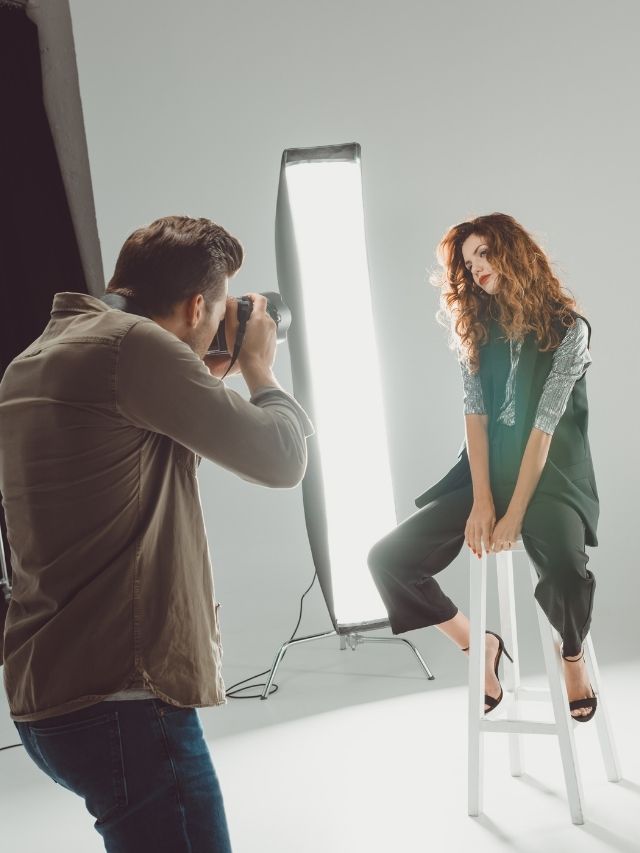 A woman is sitting on a stool while being photographed by a photographer.