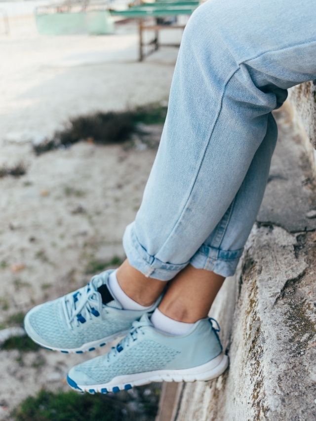 A woman's feet sitting on a wall wearing blue sneakers.