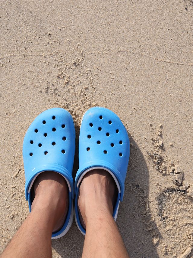 A person wearing blue crocs in the sand.