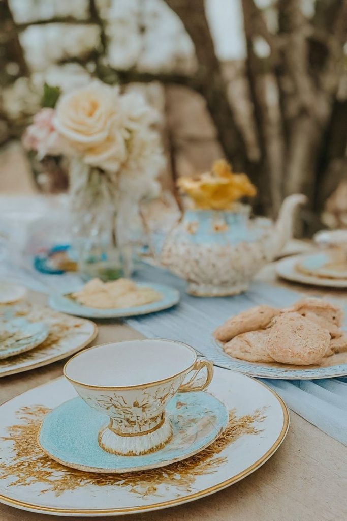 An elegant tea setting with a vintage teacup, teapot, plates, and cookies on an outdoor table, accompanied by floral decorations.