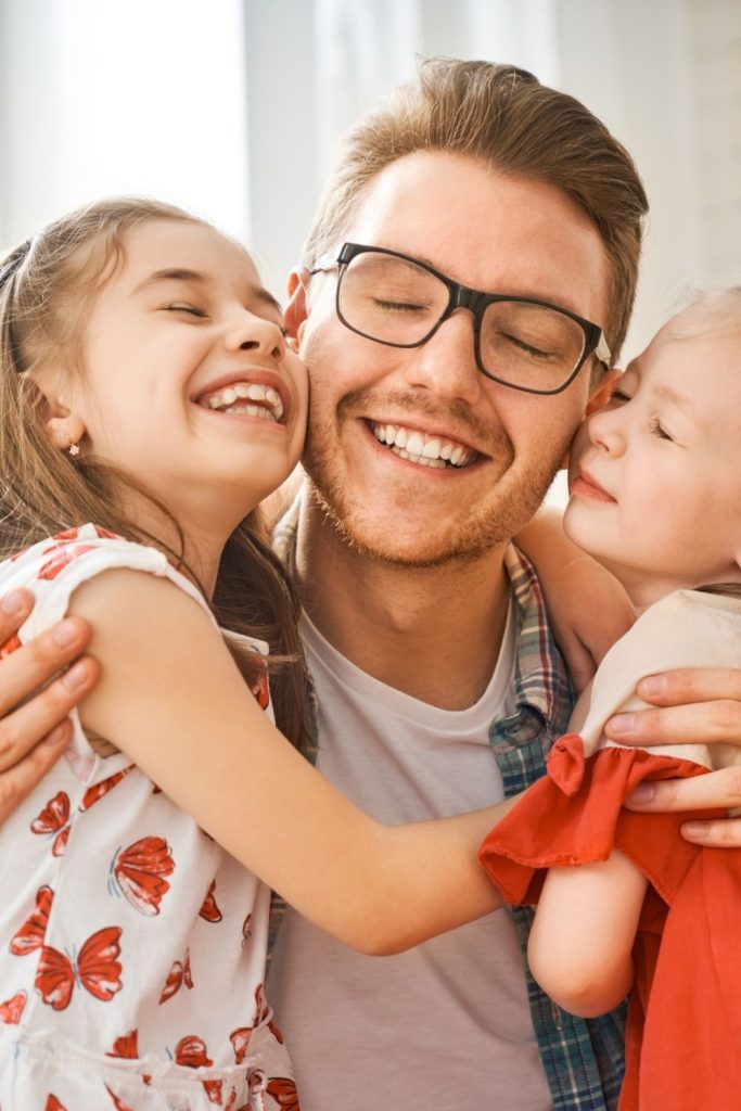 A smiling man with glasses embraces two young children who are laughing and showing affection towards him.